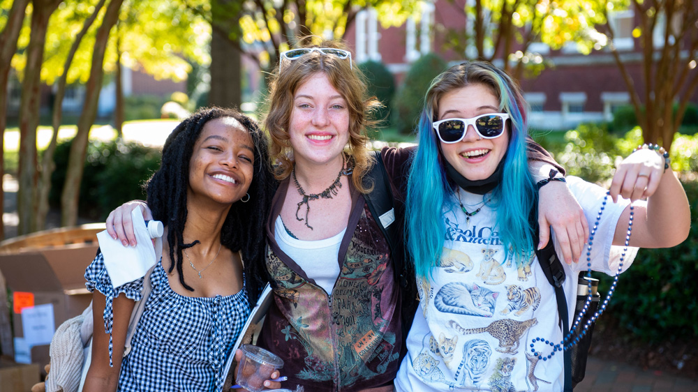 Three students, with arms around one another, share a laugh on campus.