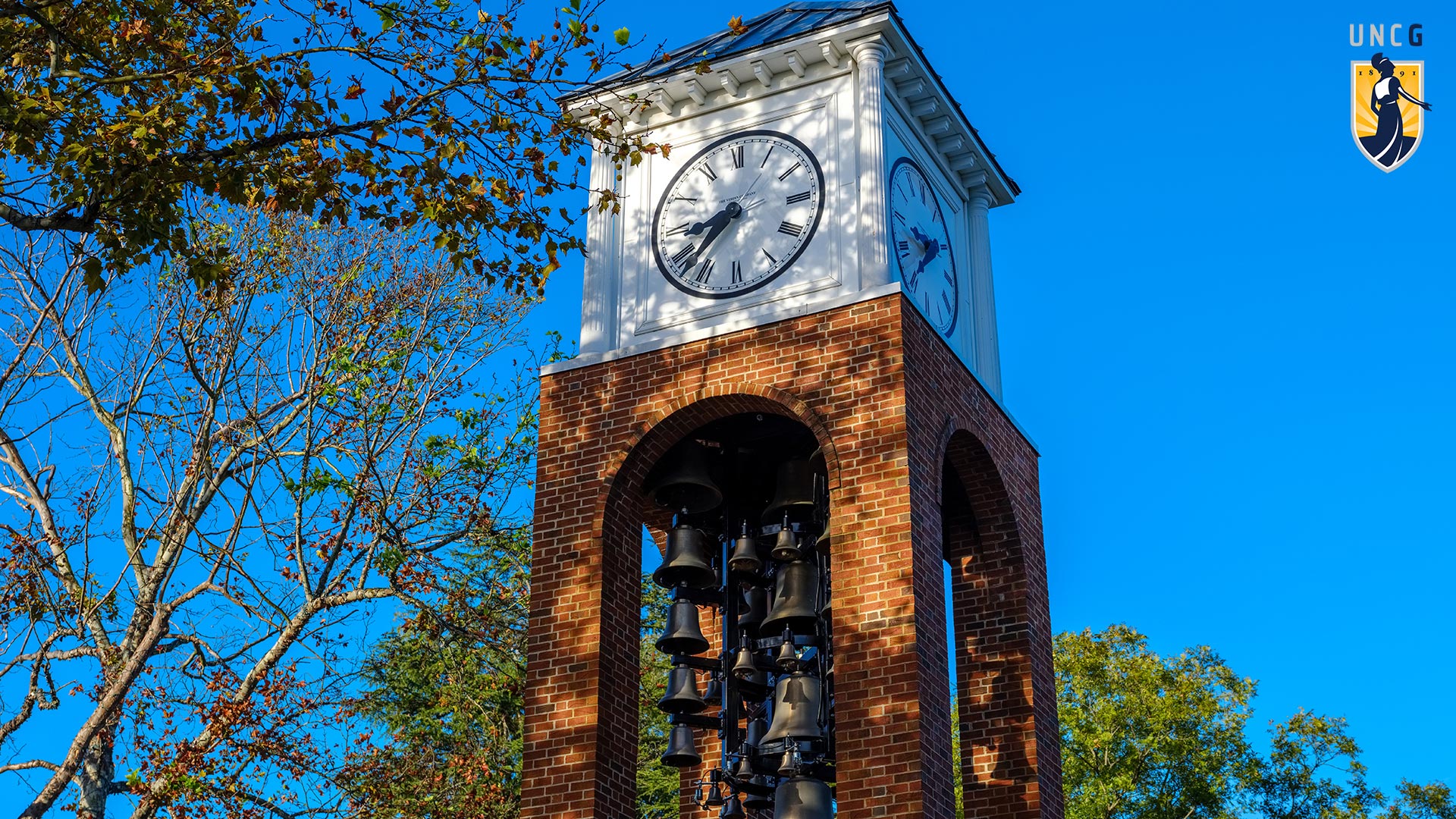 The UNCG Bell Tower against a blue sky with the UNCG logo in the top right corner