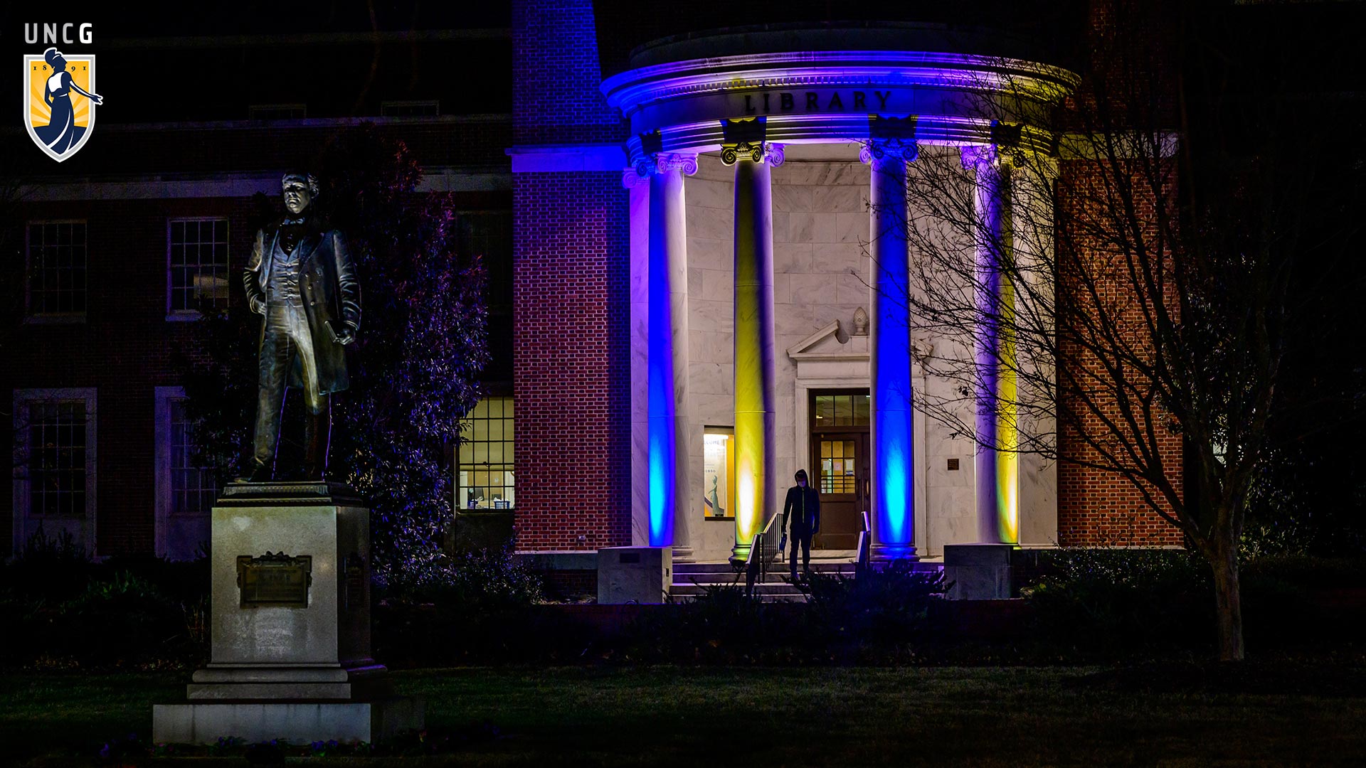 Jackson Library lit at night showing the McIver Statue in the foreground with the UNCG logo in top left corner