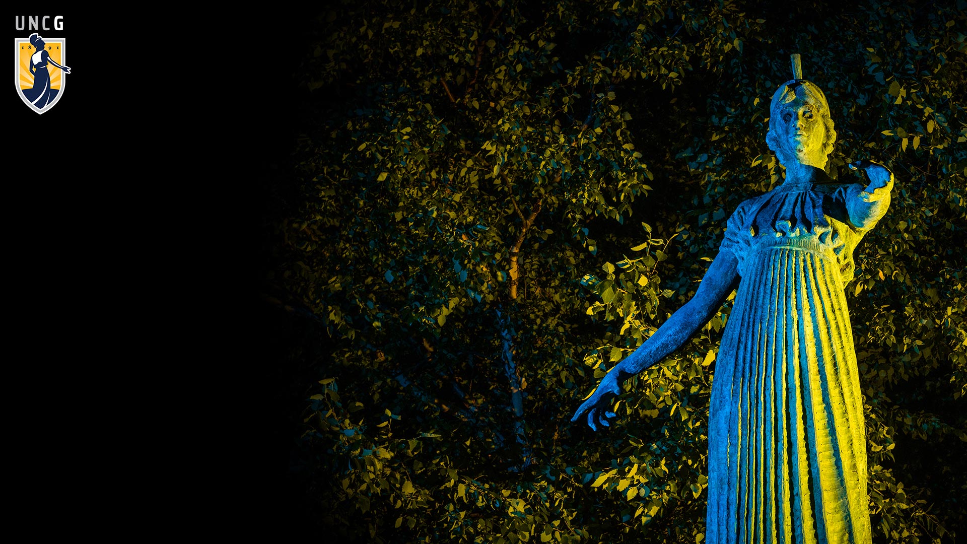Minerva Statue in front of a tree against the night sky and the UNCG logo in top left corner