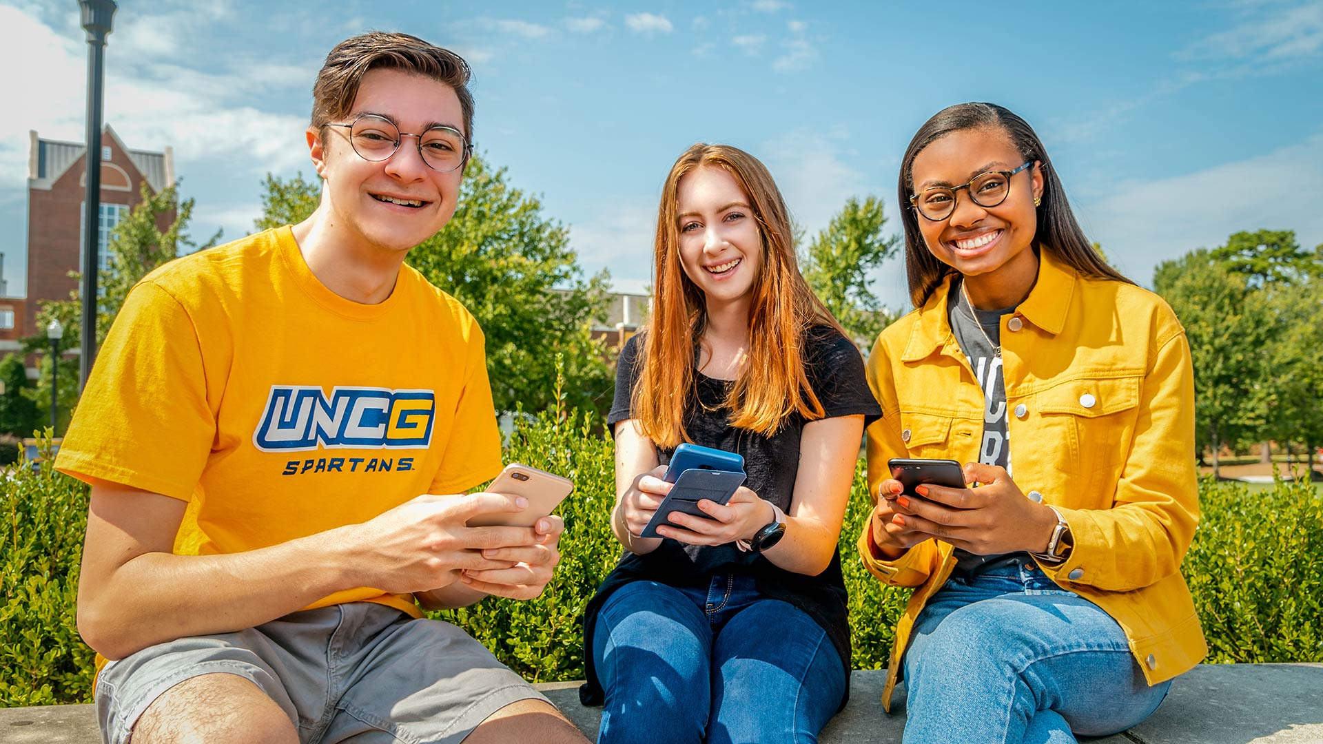 Three students seated outside smile as they hold cell phones in their hands.
