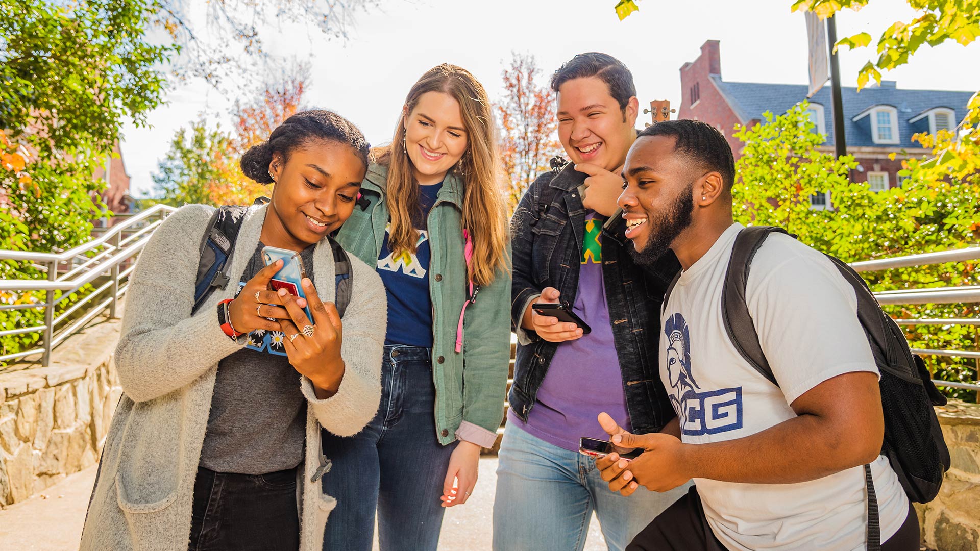 Four students smile as they look at a cell phone screen while standing outside on the UNCG campus.