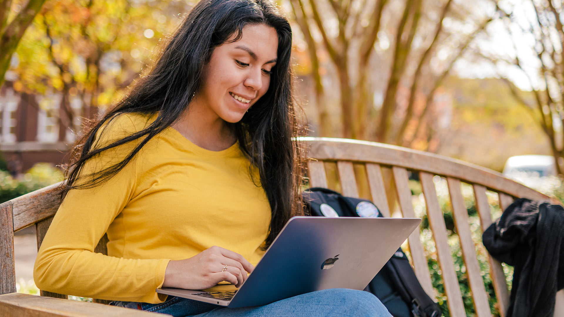 A student seated on an outdoor bench smiles as she views a laptop screen.