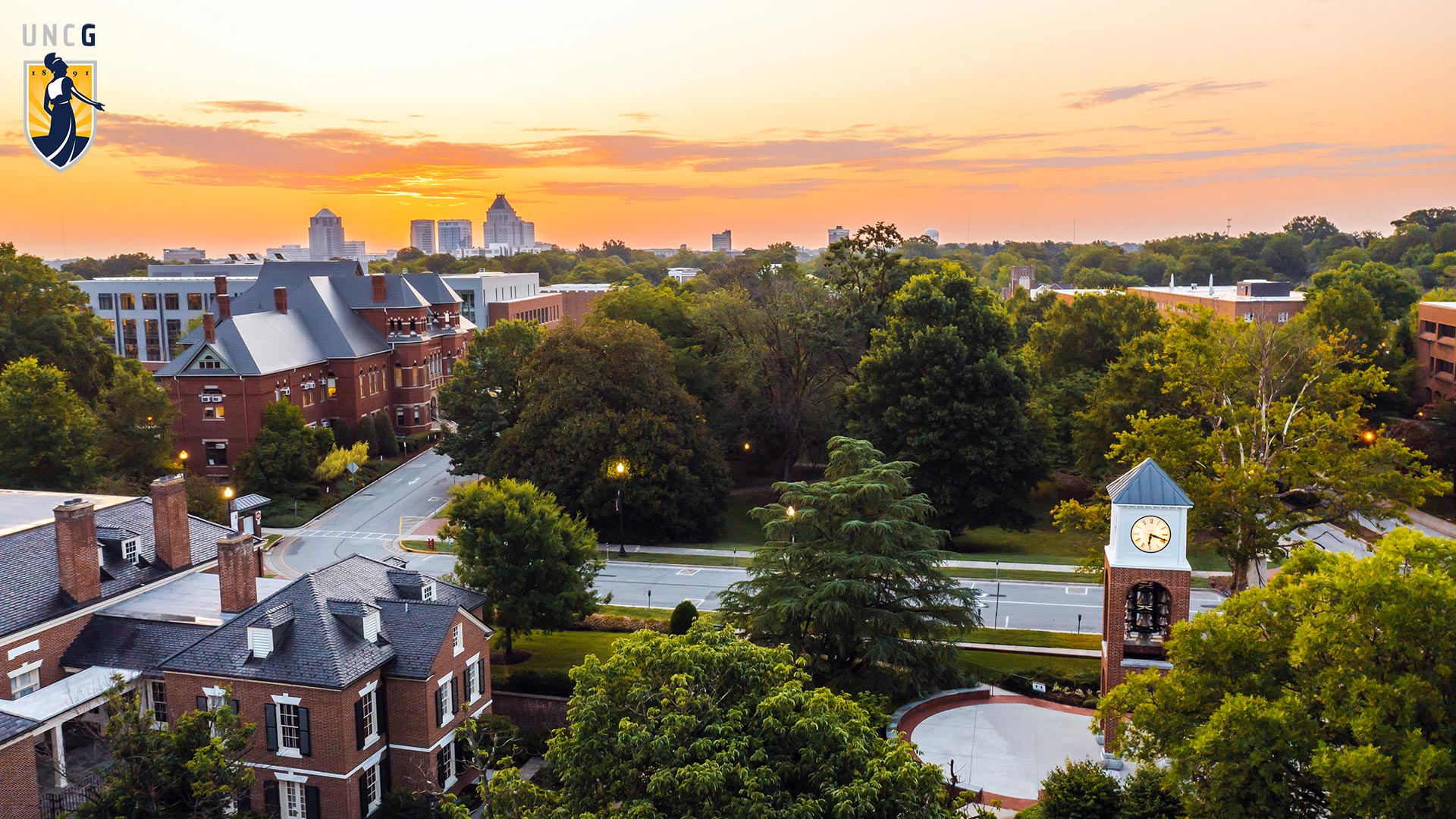 Aerial view over campus showing a sunset in the summer with the UNCG logo in the top left corner
